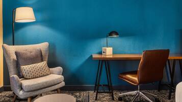 Ein Zimmer mit blauer Wand, einem grauen Sessel mit gemustertem Kissen, einem Holztisch und einem braunen Ledersessel, sowie einer Stehlampe.