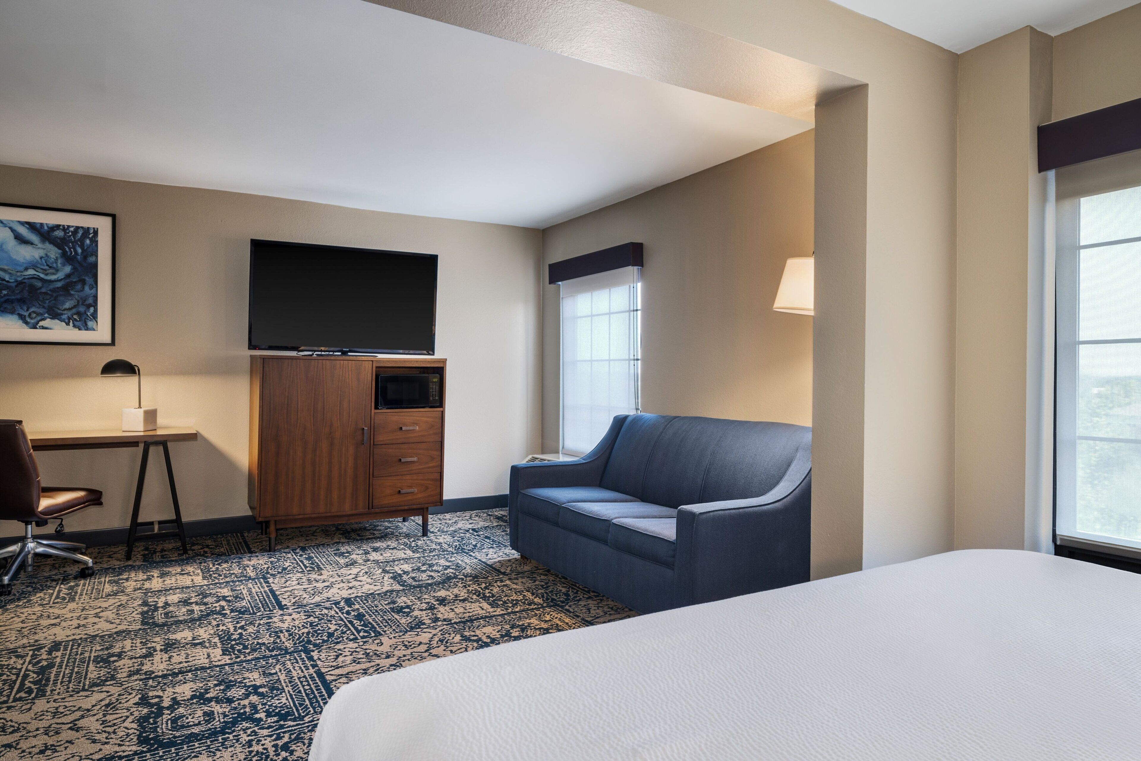 A hotel room with a large bed, a desk with a chair, a television, a sofa, and a window with a view.