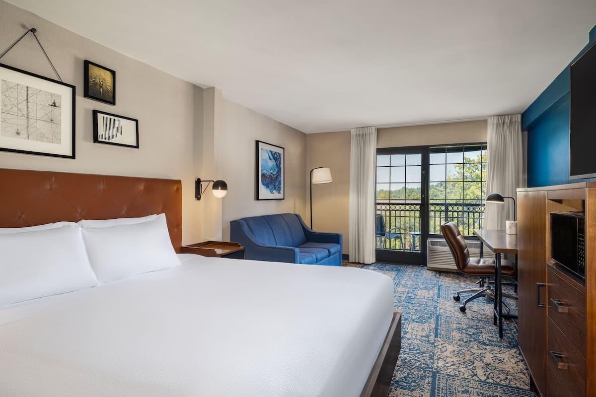 A hotel room with a large bed, a desk, a chair, and a view of the outdoors through a sliding door.