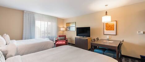 A hotel room with two beds, a TV, a desk with chairs, and a window with curtains.