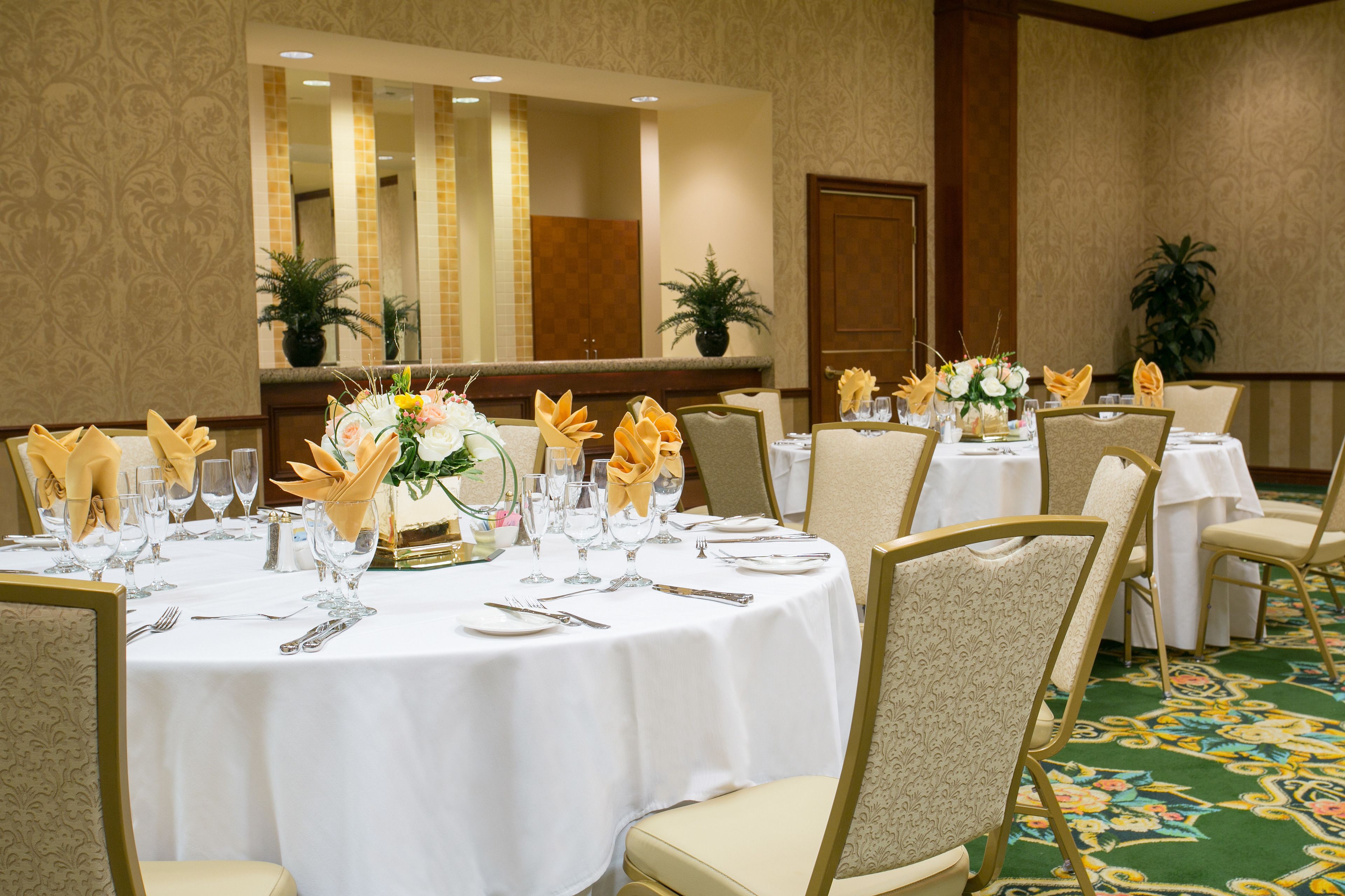 Une salle de banquet avec des tables rondes dressées pour le repas, chacune étant recouverte d’une nappe blanche, agrémentée de chaises dorées et d’arrangements floraux.