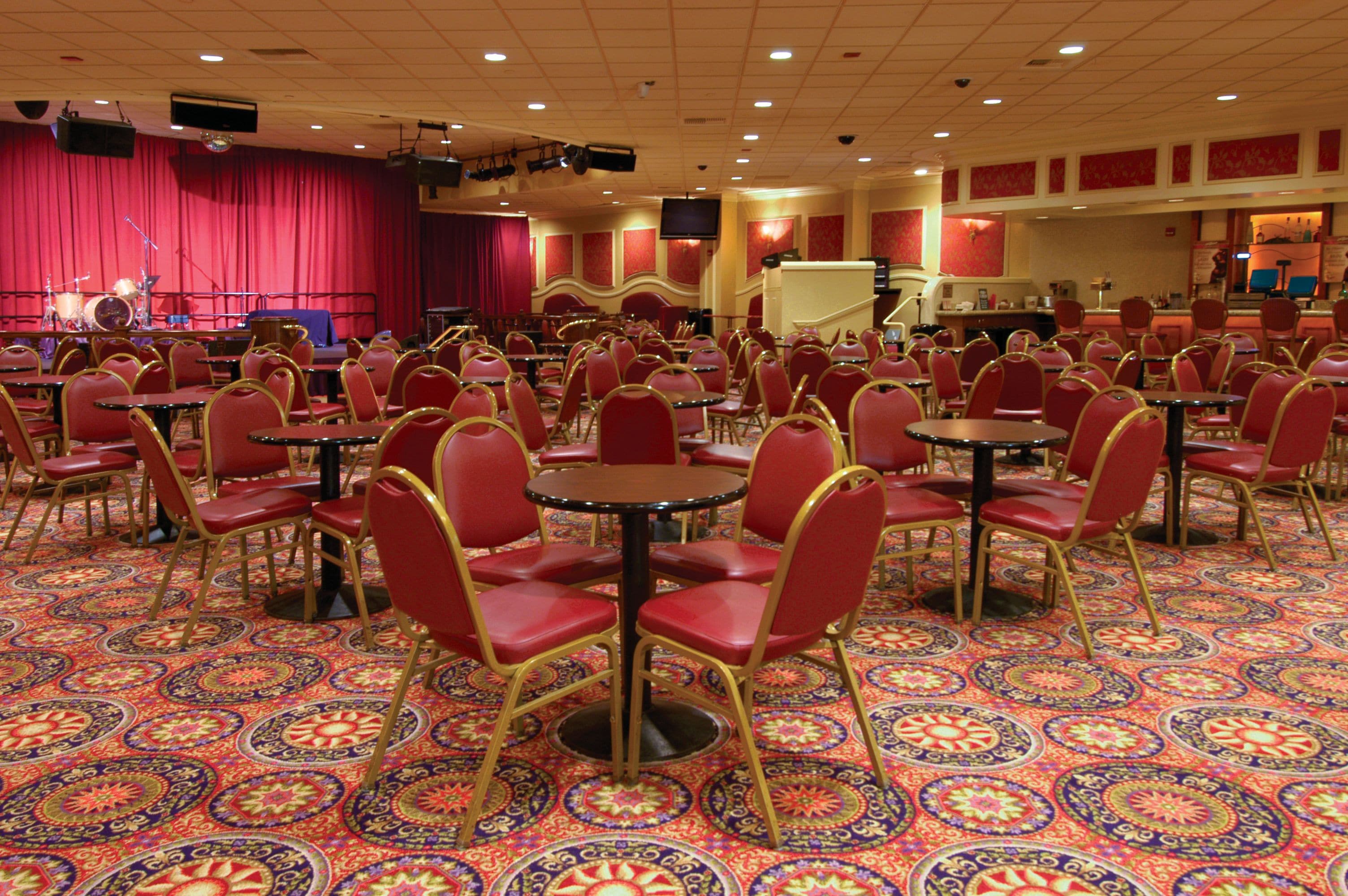A banquet hall with round tables and red chairs, a stage with musical instruments, and a patterned carpet.