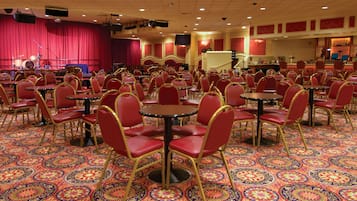 A banquet hall with round tables and red chairs, a stage with musical instruments, and a patterned carpet.