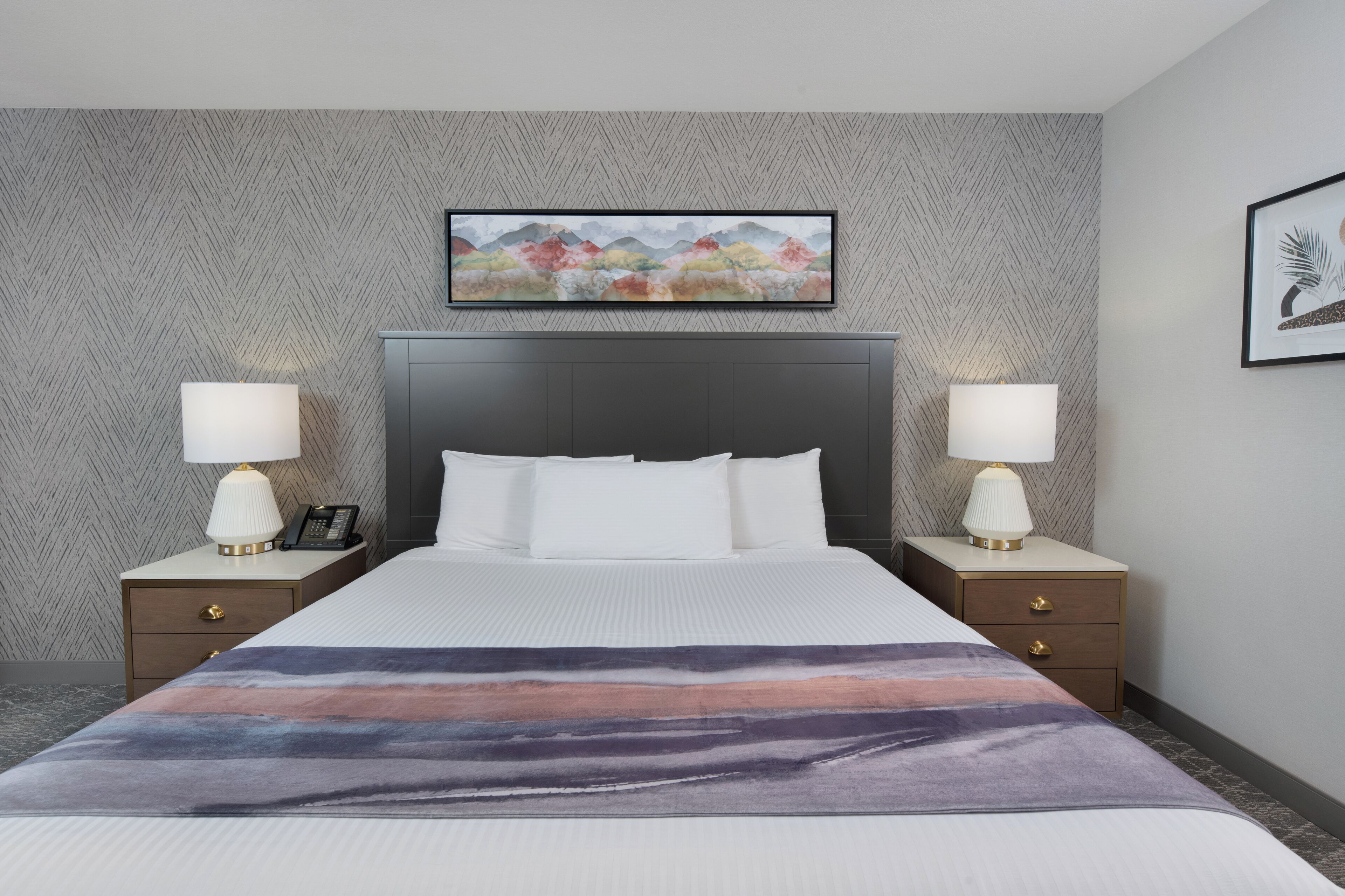 Une chambre d’hôtel avec un grand lit, deux tables de chevet avec des lampes, une œuvre d’art encadrée au-dessus du lit et un mur orné d’un motif en chevrons.