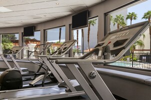 Un gimnasio con cintas de correr y equipo de ejercicio, ventanas amplias con vistas a palmeras y una piscina.