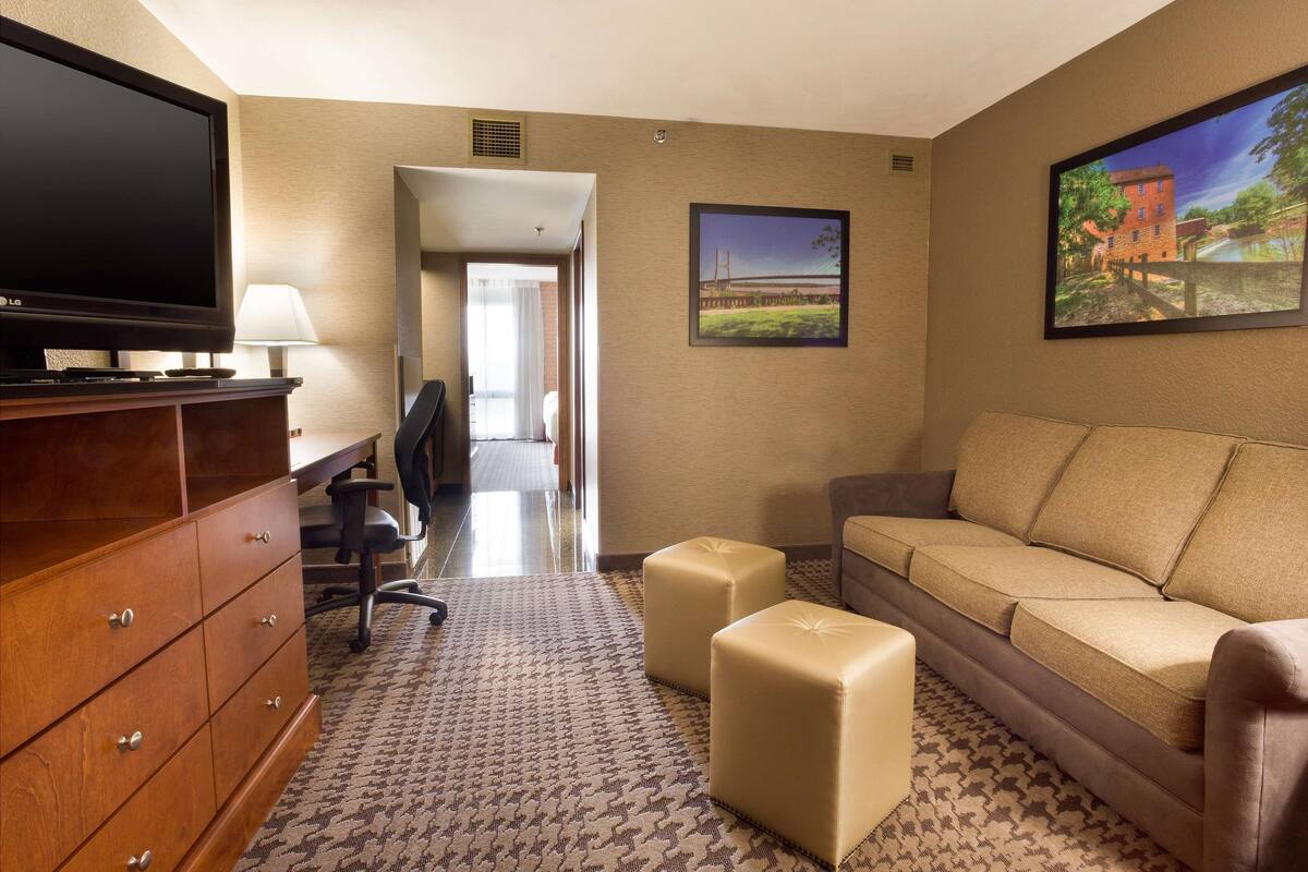 A hotel room with a flat-screen TV, wooden dresser, beige sofa, two ottomans, and a patterned rug.