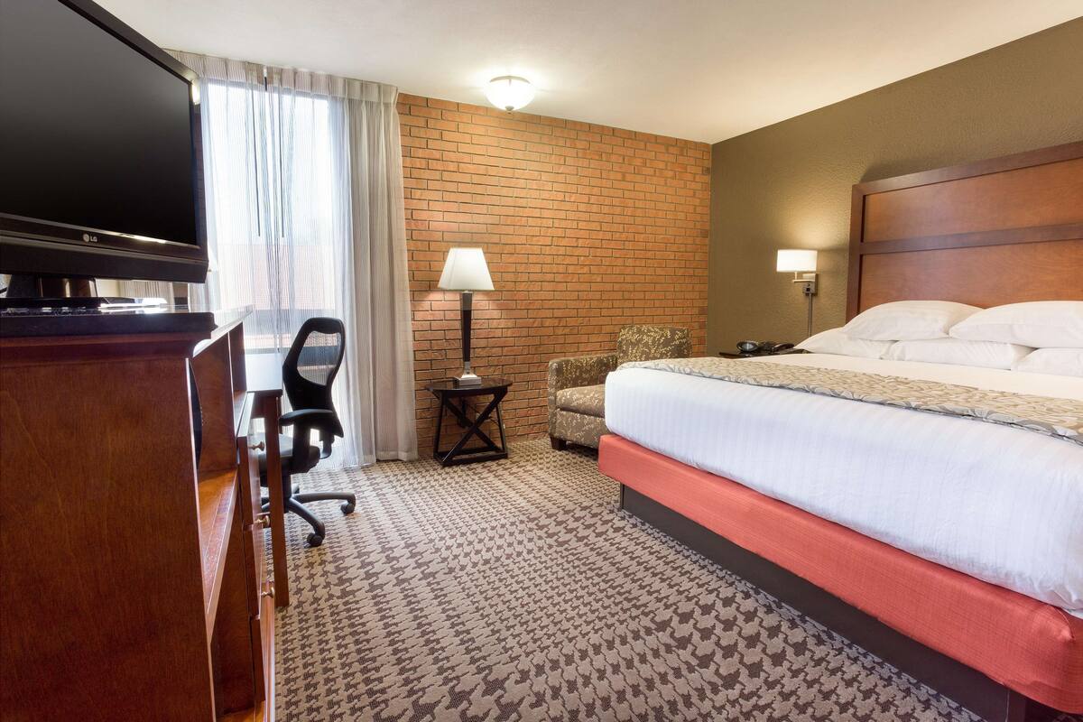 A hotel room with a large bed, a desk with a flat-screen TV, a chair, a lamp, and a brick-patterned wall.