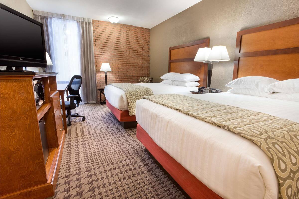 A hotel room with two beds, a TV, a desk with a lamp, and a brick wall.