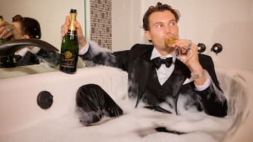 A person in a bathtub filled with bubbles, holding a bottle of champagne.