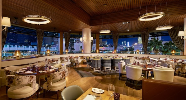 Un restaurant offrant une vue sur les toits d’une ville illuminée la nuit, avec un plafond en bois, des tables rondes et un éclairage moderne.