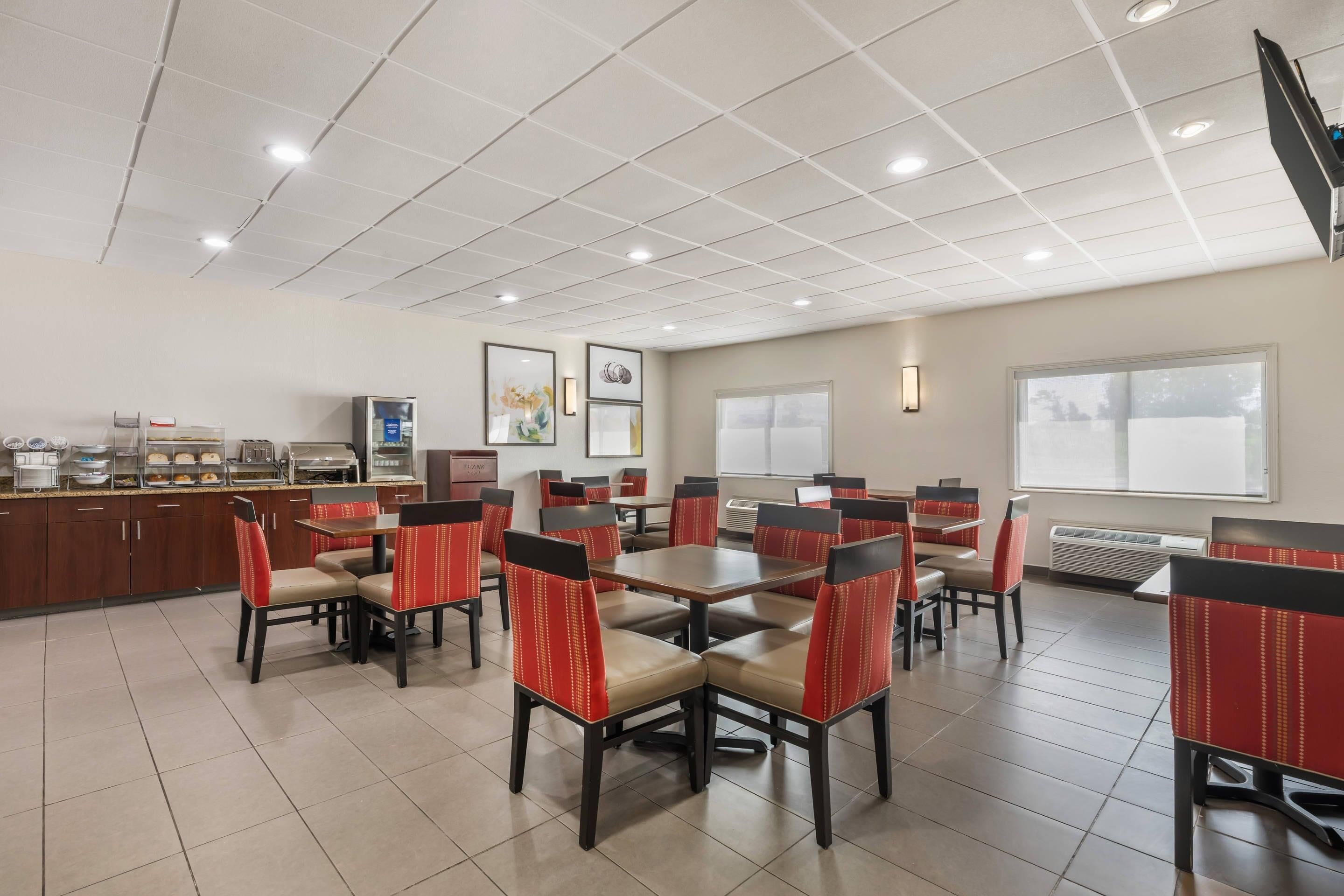 Un espace de restauration d’hôtel comprenant plusieurs tables et chaises, un coin cuisine équipé d’appareils électroménagers et de grandes fenêtres laissant entrer la lumière naturelle.