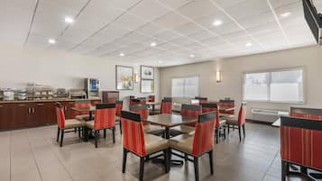Un espace de restauration d’hôtel comprenant plusieurs tables et chaises, un coin cuisine équipé d’appareils électroménagers et de grandes fenêtres laissant entrer la lumière naturelle.