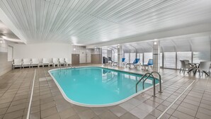 Una piscina interior con suelo de baldosas, una escalera de piscina y sillas y mesas blancas alrededor del área de la piscina.