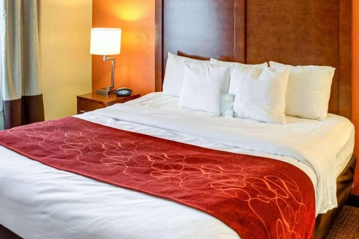 A hotel room with a large bed, a wooden headboard, a nightstand with a lamp, and a red throw blanket.