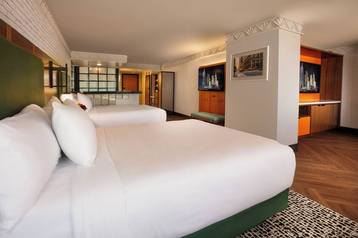 Ein Hotelzimmer mit zwei großen Betten, einer kleinen Küchenzeile und einer Sitzecke mit Blick auf die Stadtlandschaft.