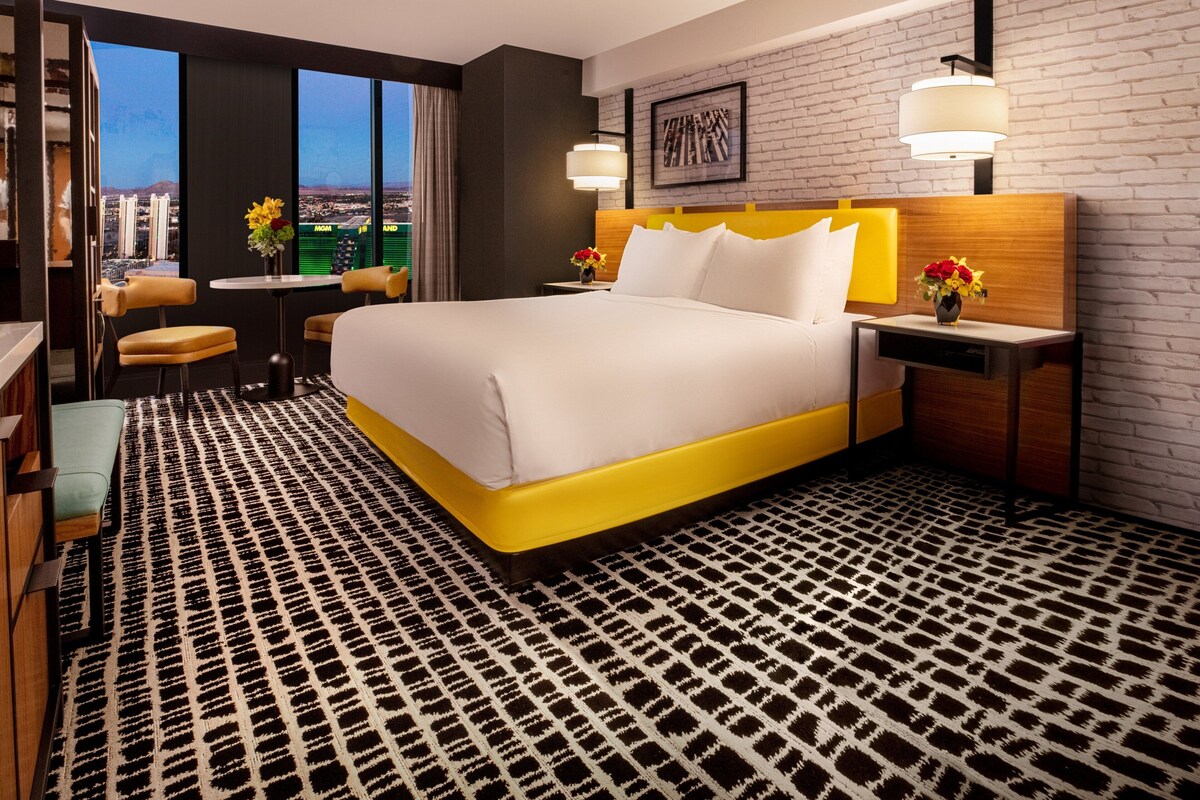 A modern hotel room with a large bed, a desk, a chair, and a view of the city.