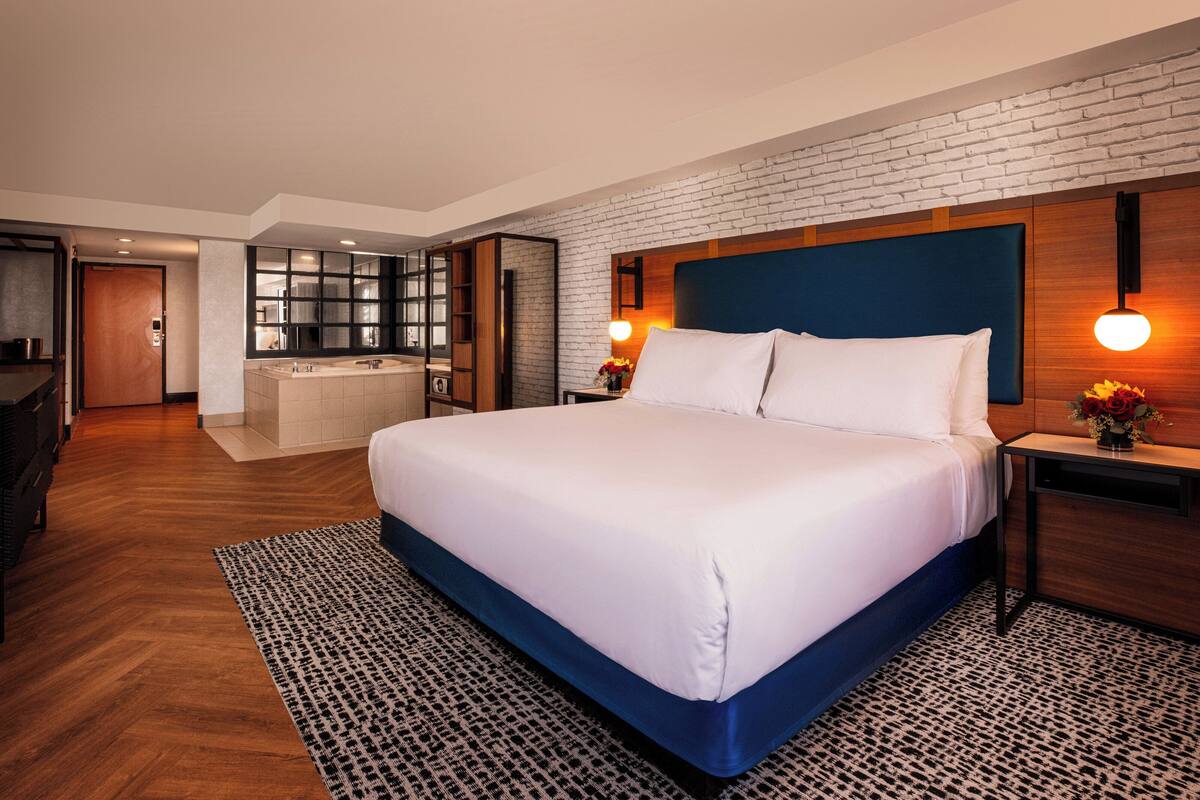 Ein modernes Hotelzimmer mit einem großen Bett, einem Holzkopfteil, einem integrierten Schreibtisch, einem Schminktisch mit Spiegel und einer Wand mit Backsteinverkleidung.