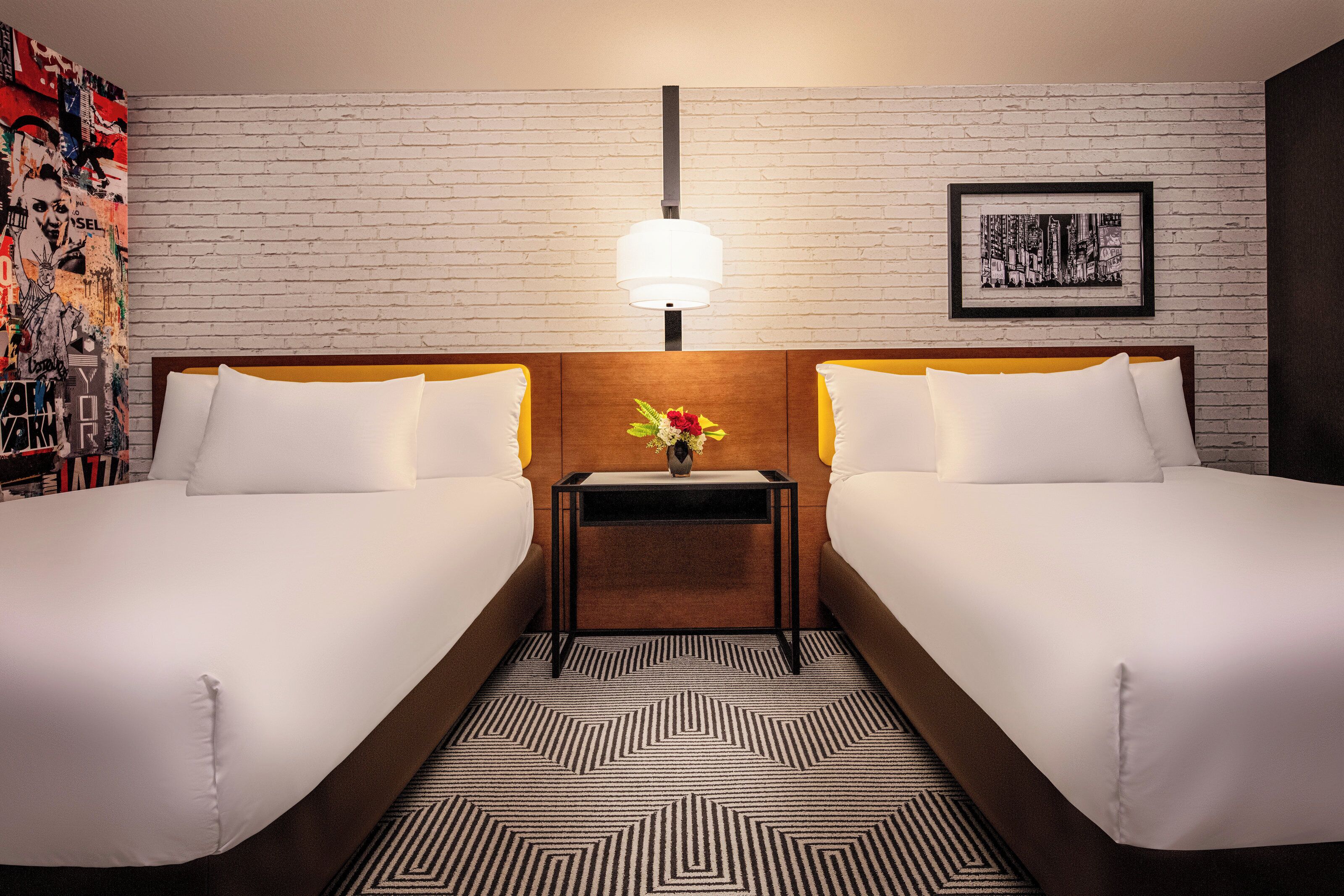 A hotel room with two beds, a nightstand with a lamp, a framed picture on the wall, and a patterned rug.