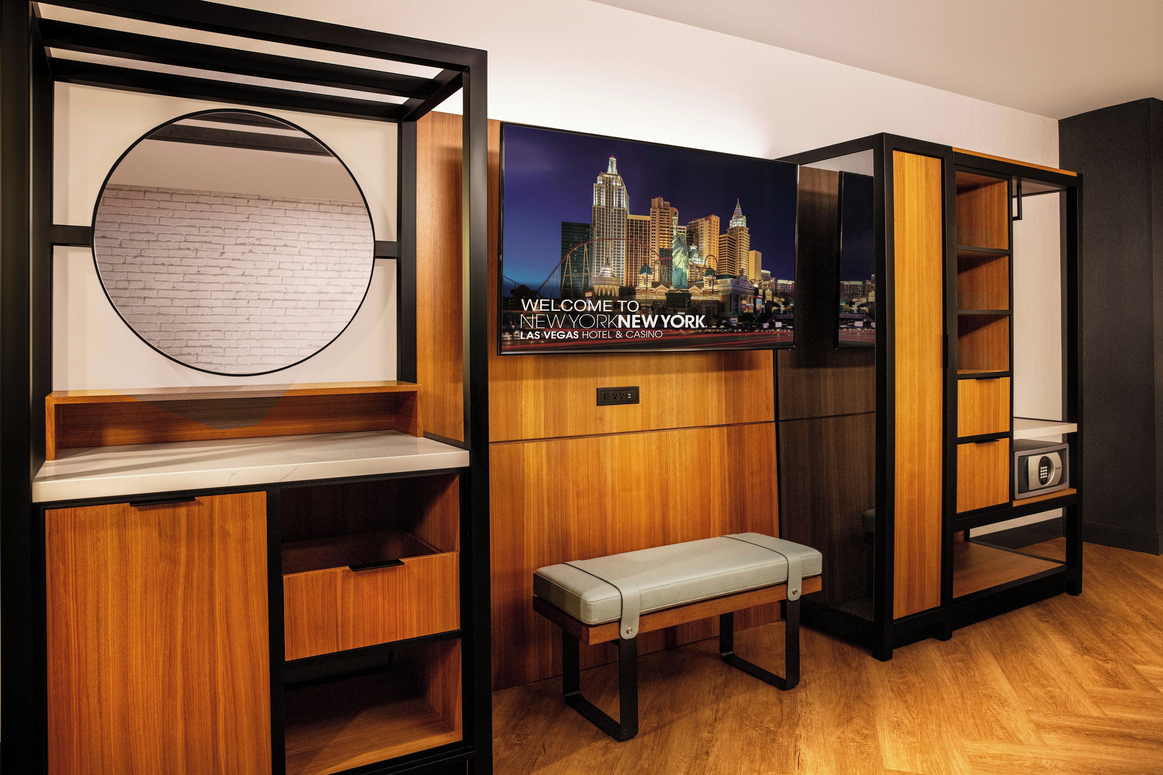 Une chambre d’hôtel avec un banc en bois, un miroir rond et un téléviseur fixé au mur qui diffuse des images d’un paysage urbain.