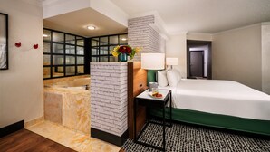 Ein Hotelzimmer mit einem großen Bett, einem Badezimmer mit einer Badewanne, einem kleinen Tisch mit einer Lampe und einer Vase mit Blumen.