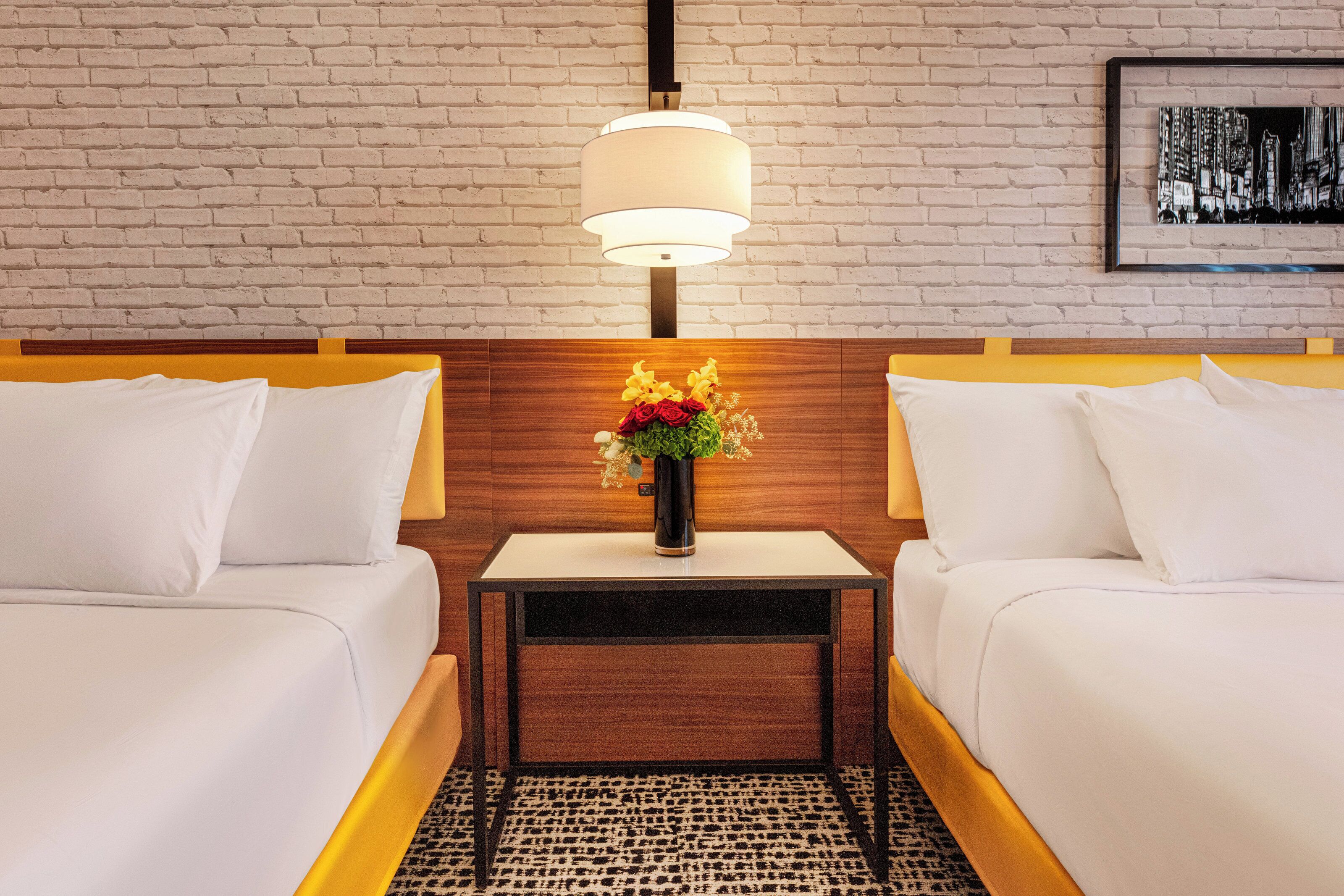 Habitación de hotel con dos camas, una mesita de noche con un florero, una lámpara y un cuadro en la pared.