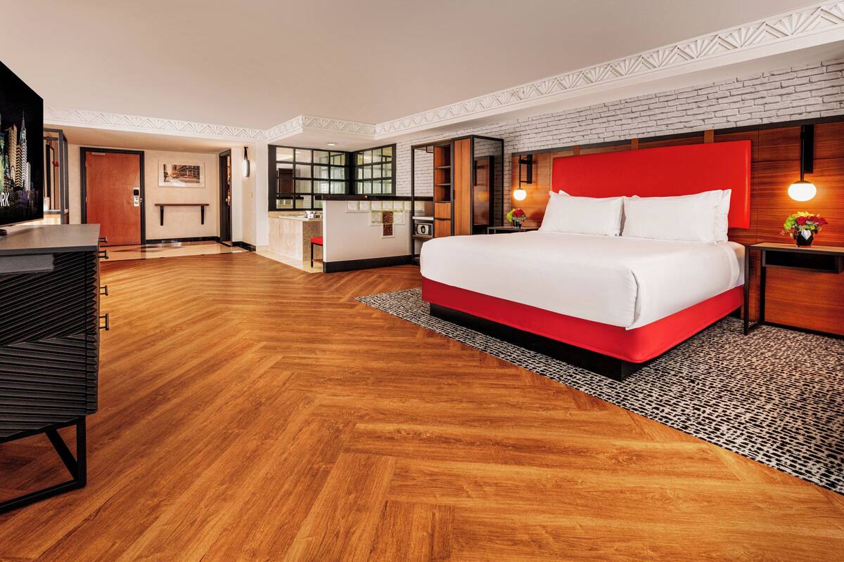 Ein modernes Hotelzimmer mit einem großen Bett, Holzboden, einer roten Akzentwand und einer Küche im Hintergrund.