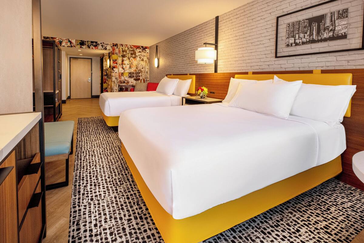 Ein Hotelzimmer mit zwei Betten, einem Schreibtisch, einem Stuhl, einem gemusterten Teppich und einer Stadtlandschaft als Wandbild.