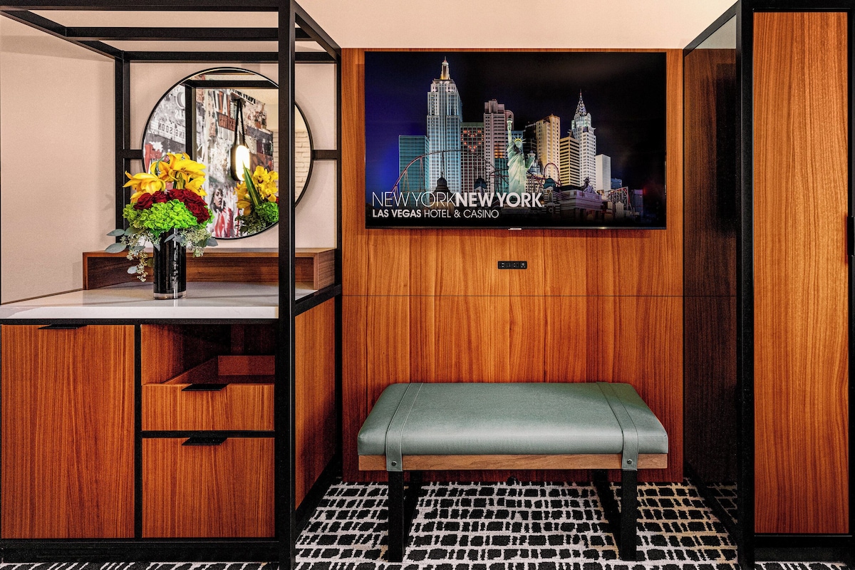 A hotel room with a wooden interior, a bench, a wall-mounted TV displaying a cityscape, and a small table with a vase of flowers.