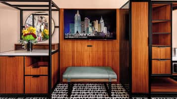 A hotel room with a wooden interior, a bench, a wall-mounted TV displaying a cityscape, and a small table with a vase of flowers.