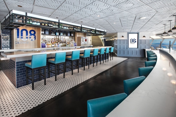 A modern bar area with a counter displaying '108 DRINKS', multiple bar stools, and a staircase leading to an upper level.