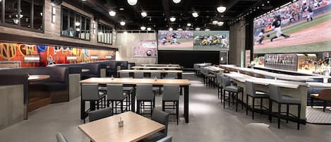 Un restaurante moderno con varias áreas de estar, incluyendo cabinas y mesas, y una barra con una pantalla grande que muestra eventos deportivos.