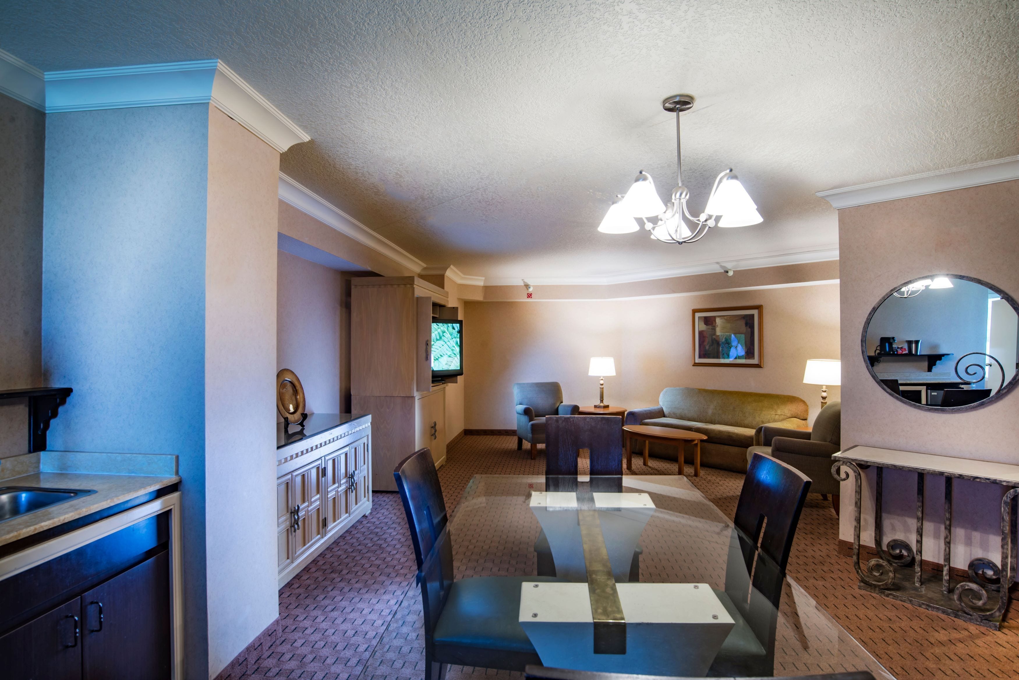 Habitación de hotel con una mesa de comedor, sillas, un sofá, un televisor, una lámpara y un espejo.