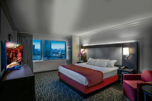 A hotel room with a large bed, a TV, a city view, and a red chair.