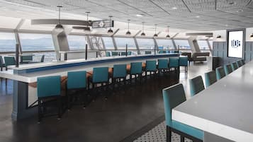 A modern bar area with a long counter, multiple seating options, and a view of the city and mountains through large windows.
