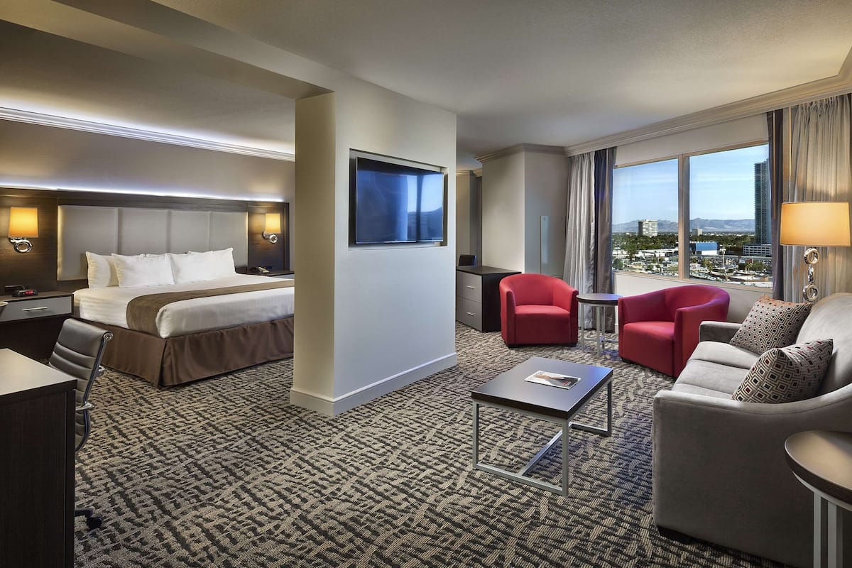 Habitación de hotel con cama, escritorio, sillón rojo, sofá gris y vista a la ciudad a través de amplias ventanas.