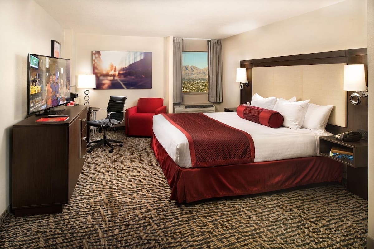 Habitación de hotel con una cama grande, un escritorio con televisor, una silla roja y una ventana con vistas a la montaña.