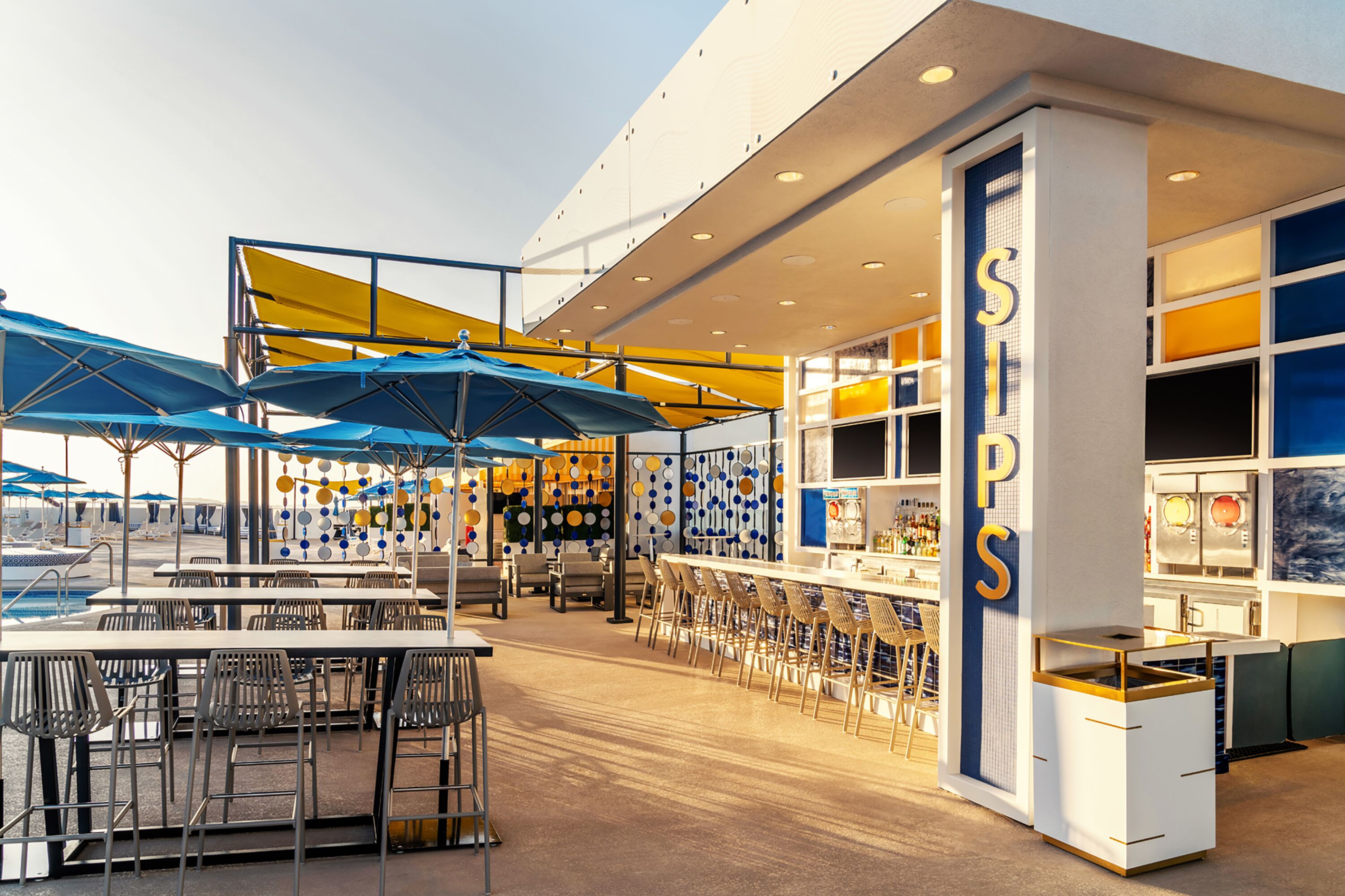 Un bar moderno con varias mesas y sillas, un área de estar junto a la piscina y una barra con un letrero que dice "SIPS".