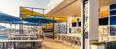 Un bar moderno con varias mesas y sillas, un área de estar junto a la piscina y una barra con un letrero que dice "SIPS".