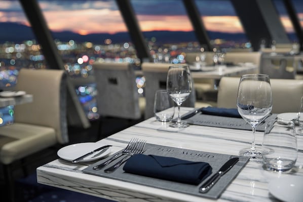 A restaurant setting with a table set for dining, a city view in the background, and a sunset over the mountains.
