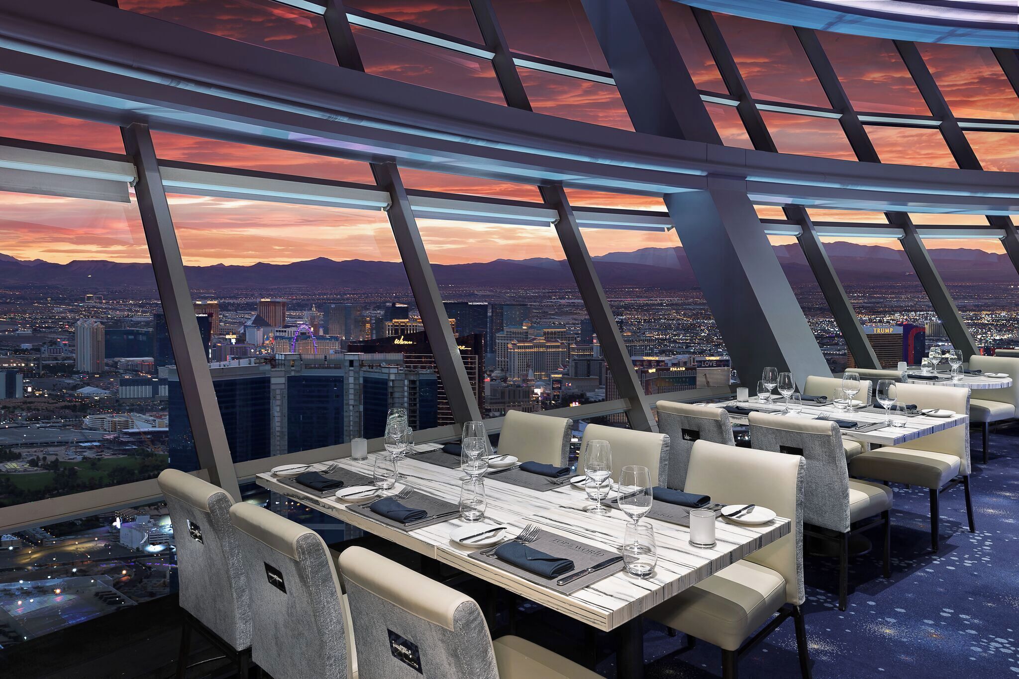 A high-rise restaurant with tables set for dining, offering a panoramic city view at sunset.