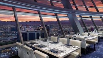 A high-rise restaurant with tables set for dining, offering a panoramic city view at sunset.