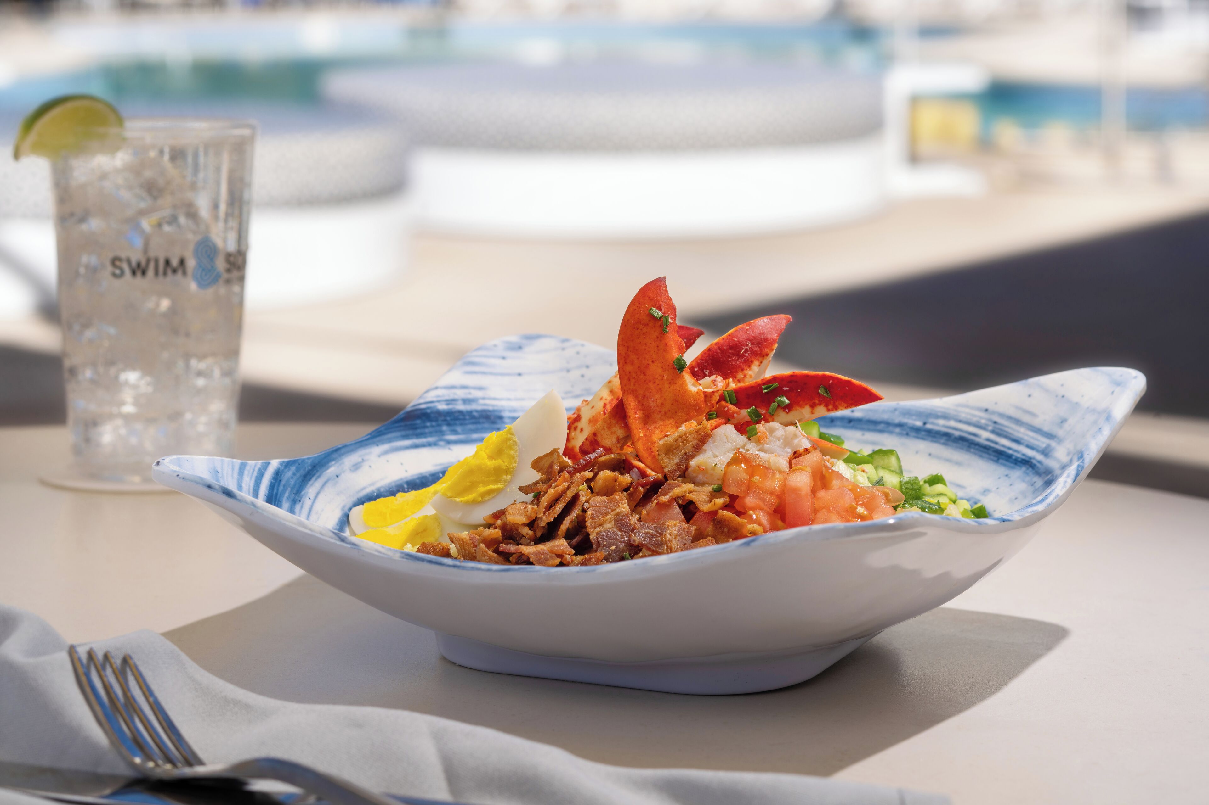 Un piatto di pesce con astice, uovo e verdure su un piatto a motivi blu e bianchi, accompagnato da un bicchiere d'acqua etichettato 