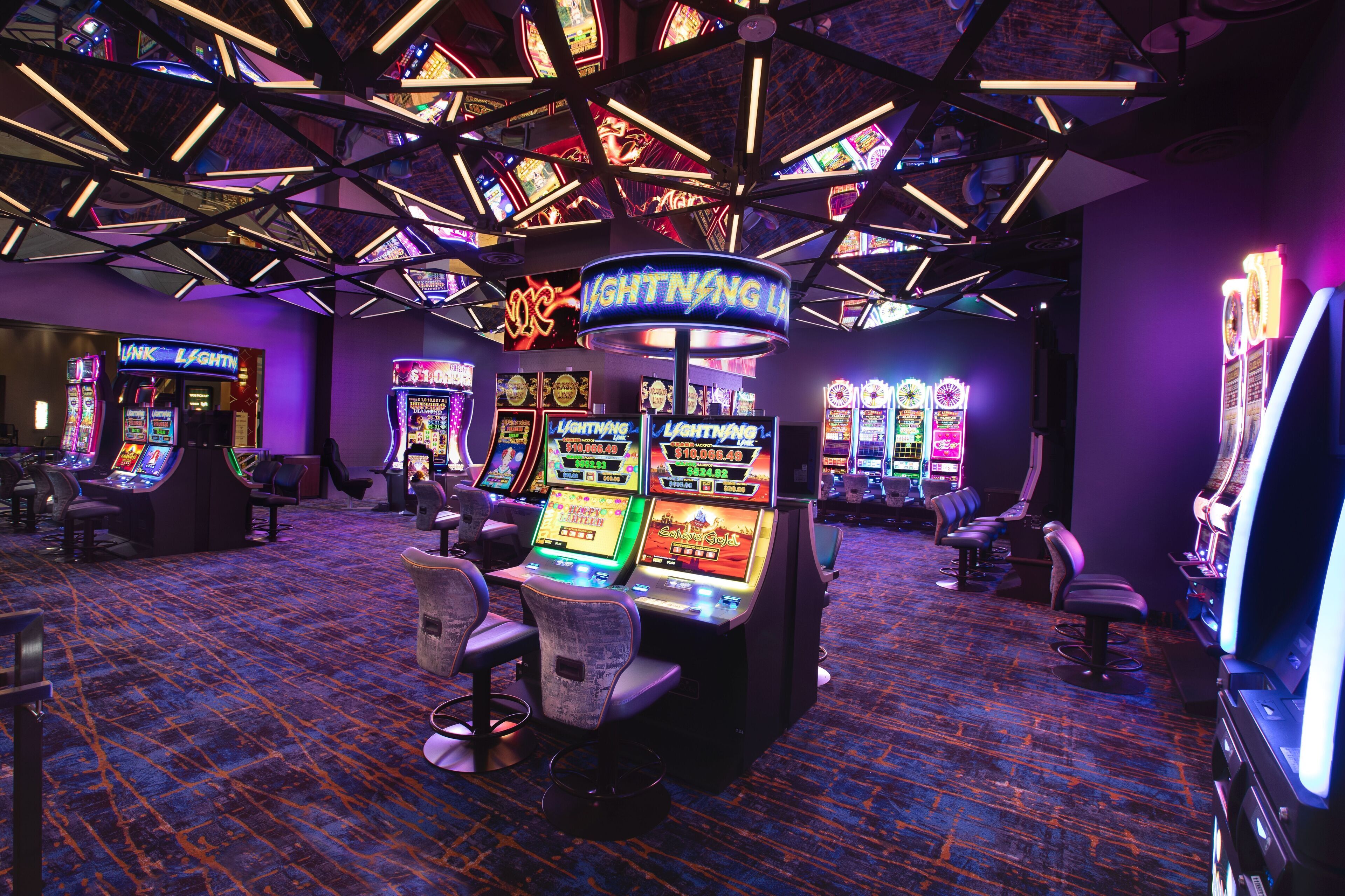 A casino with slot machines and neon signs, featuring a modern ceiling design and carpeted floor.