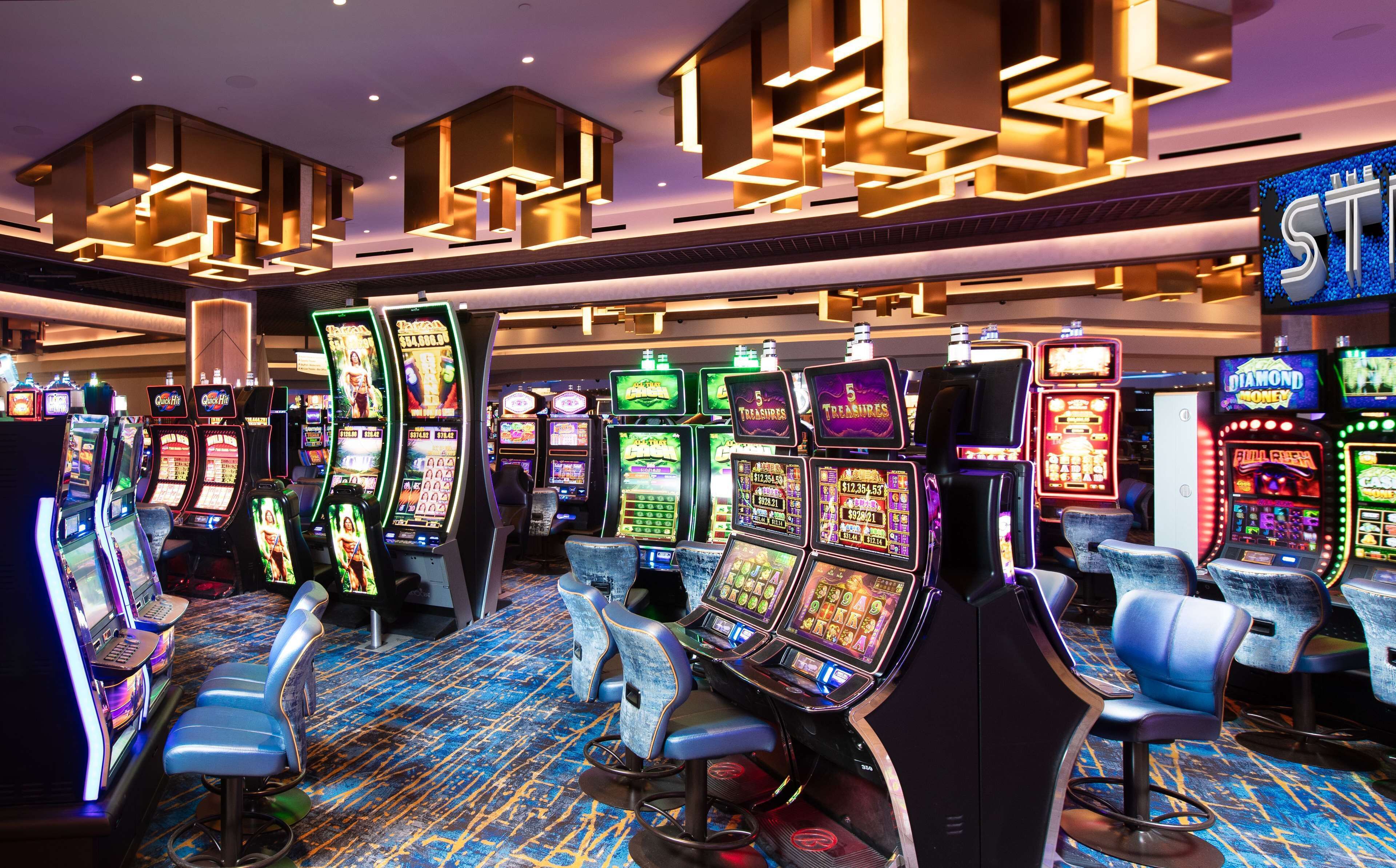 A casino floor with multiple slot machines and gaming tables, illuminated by overhead lighting.
