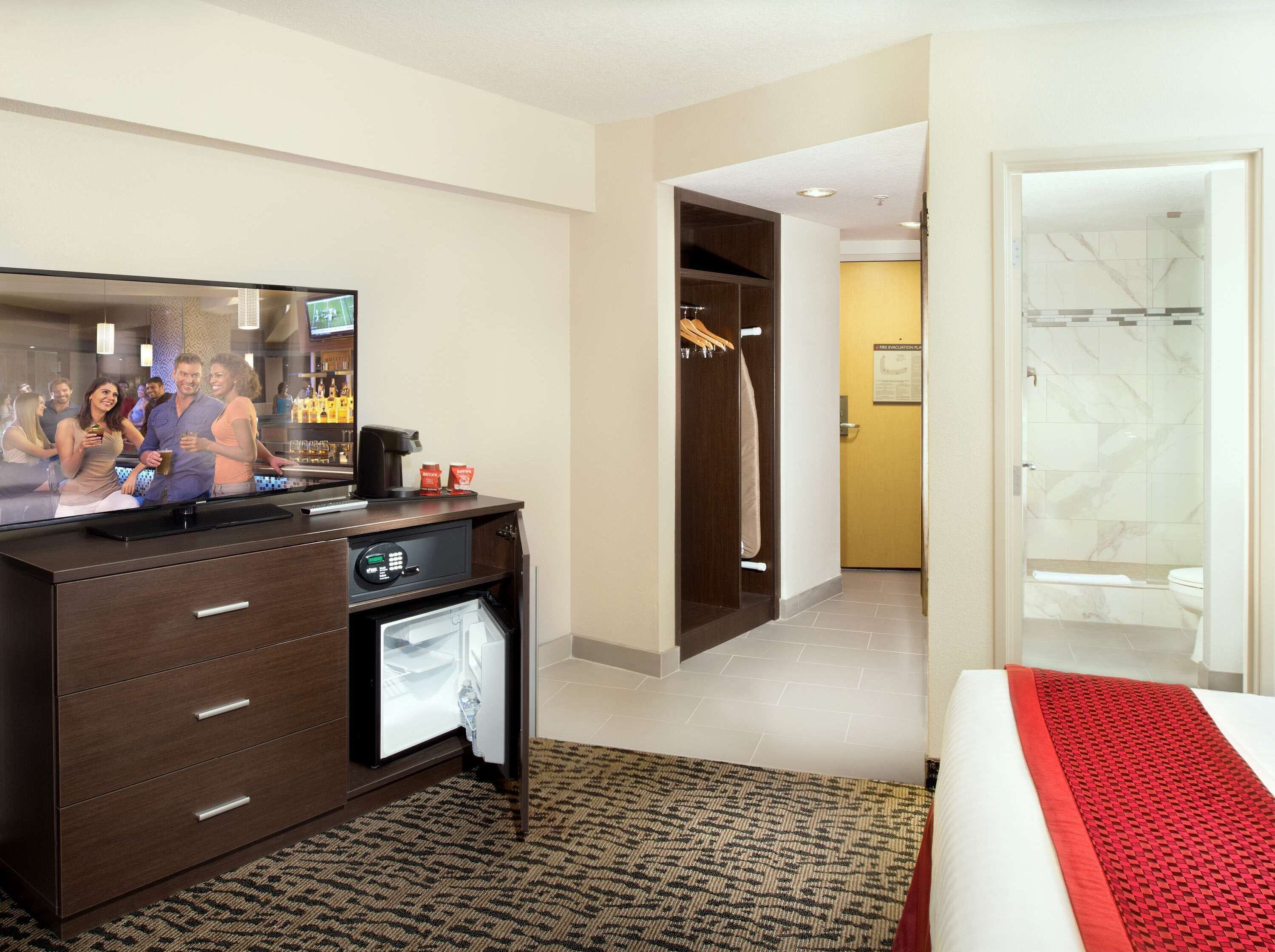 Camera d'albergo con un minibar, televisore, bagno con doccia e un letto con cuscino rosso.