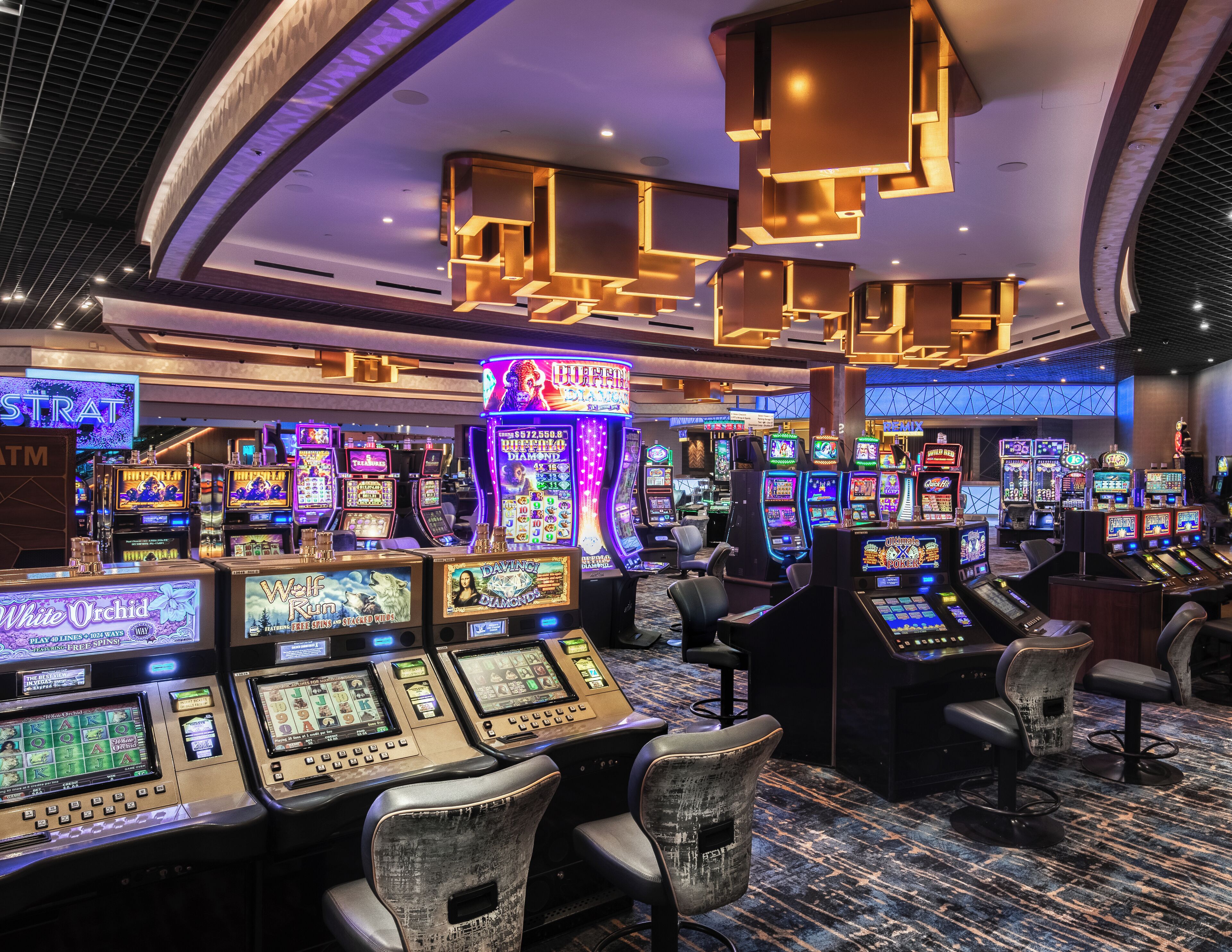 Un casino doté de nombreux appareils à sous, de tabourets de bar et d’un plafond orné de luminaires aux formes géométriques.