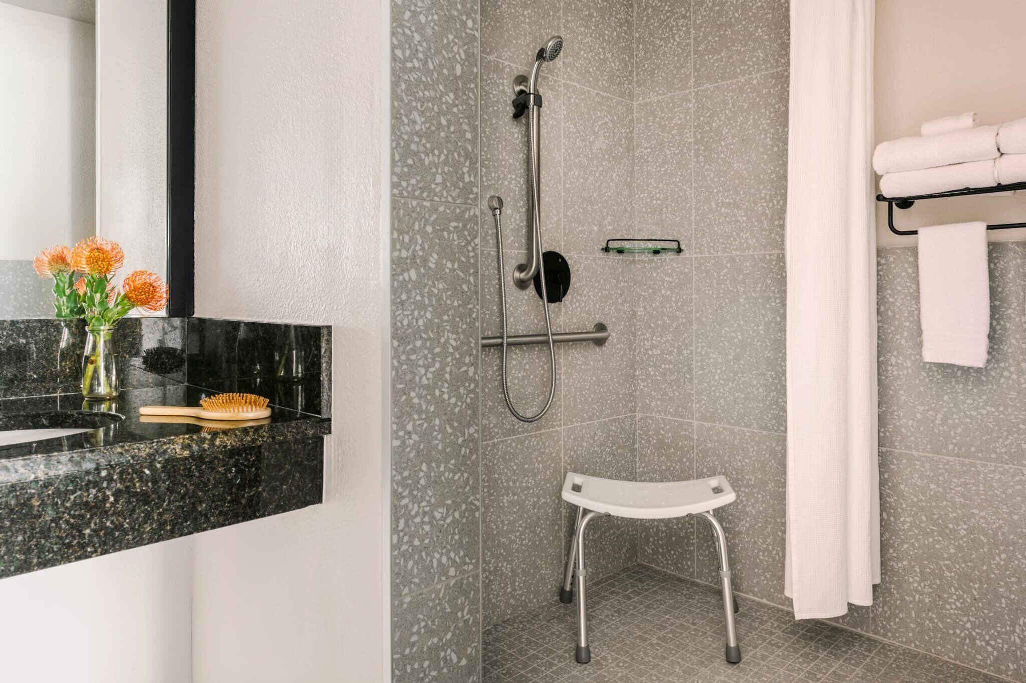 A modern bathroom with a shower, a small white stool, a towel rack, and a vase with flowers on the counter.