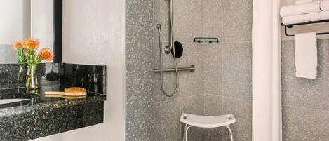 Une salle de bain moderne avec une douche, un petit tabouret blanc, un porte-serviettes et un vase avec des fleurs posé sur le comptoir.