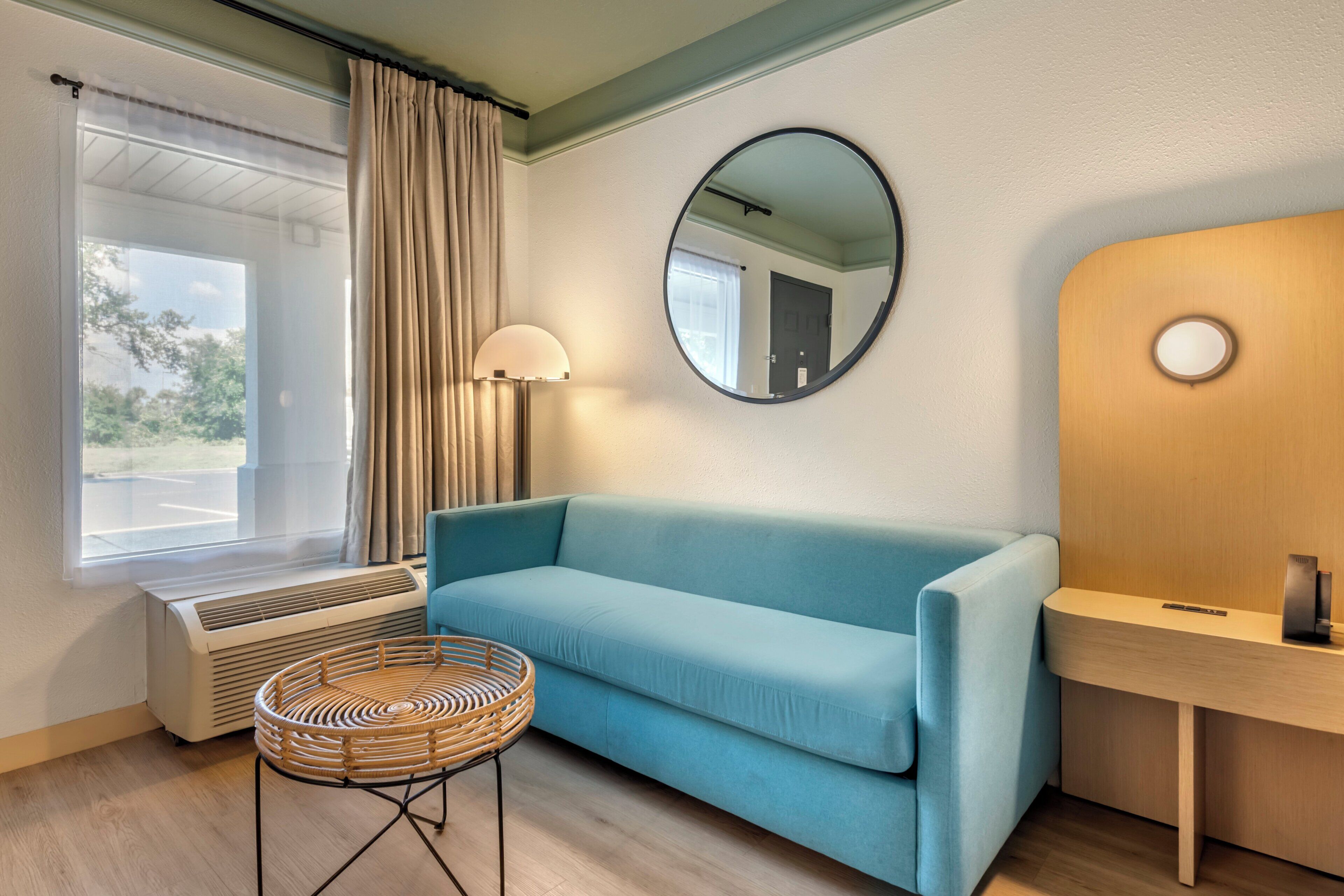 A room with a blue sofa, a round wicker table, a mirror, a lamp, and a wooden desk.