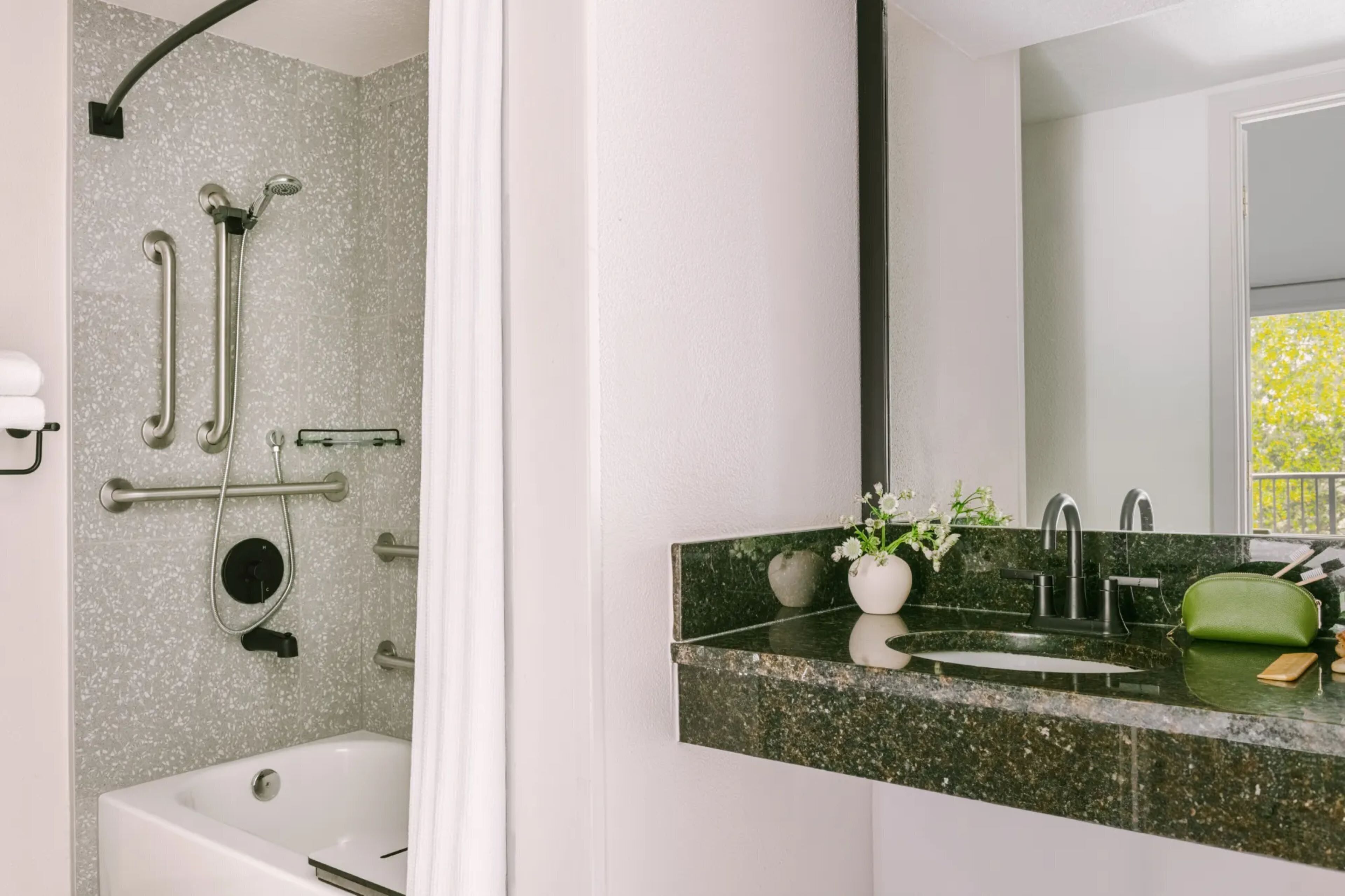 A bathroom with a shower, grab bars, a bathtub, a sink with a green bag, and a mirror.